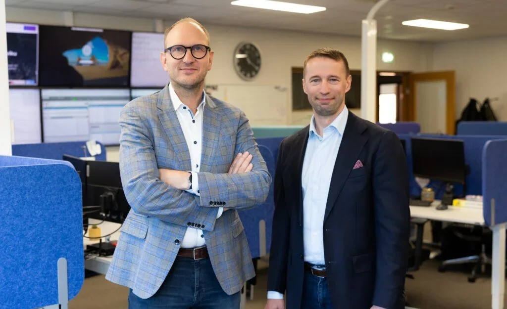 Hansab grupas izpilddirektora un Replicant IT izpilddirektora kopbilde ar biroju fonā. Abas personas smaidoši skatās kamerā.
